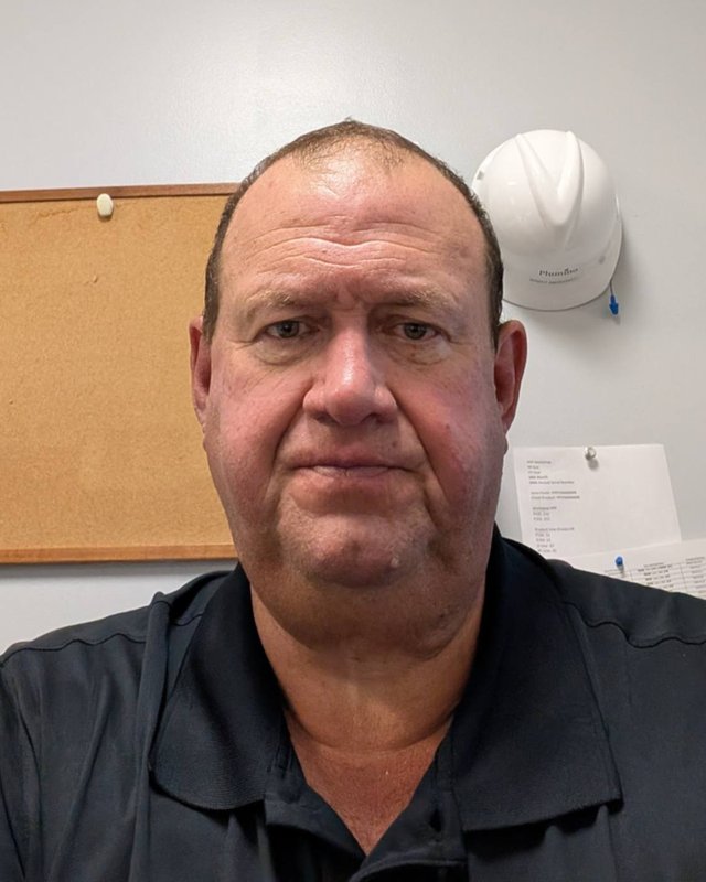 Brent Ashcraft in a black polo shirt in his office, a white hard hat hanging on the wall behind him.