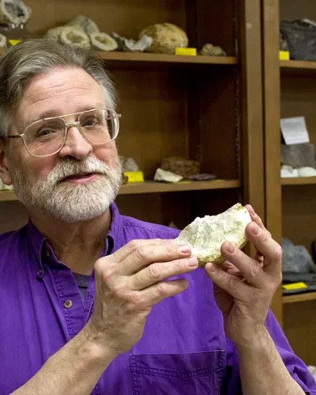 Mike Fix holding a rock specimen in front of shelves of labelled rocks and minerals.