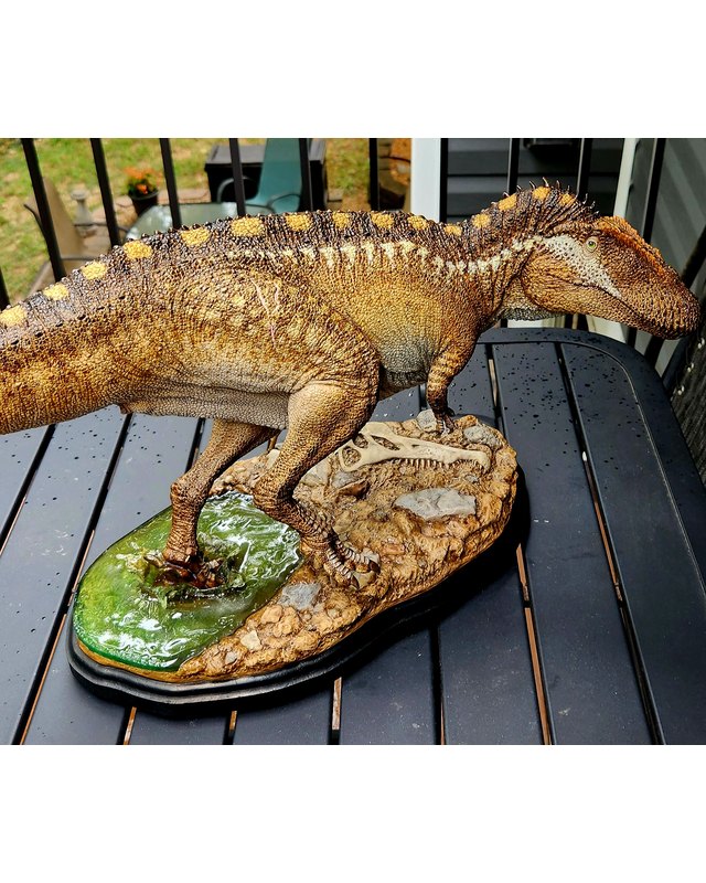 A sculpture of a large theropod with a ridged back striped in gold and brown, stepping from a pool onto rock beside a buried skull, by Shane Foulkes.