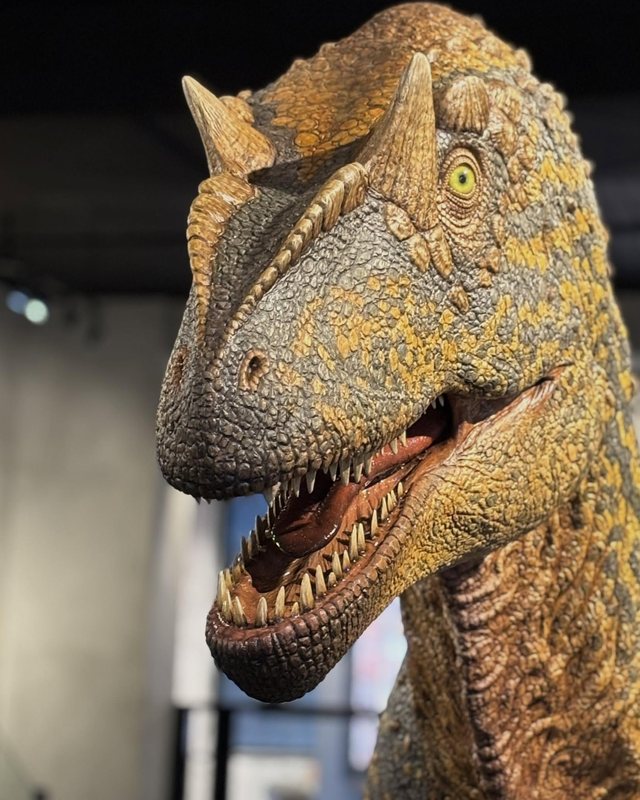 A life size Allosaurus sculpture with mottled gold and grey skin, head turned toward the viewer, by Shane Foulkes.
