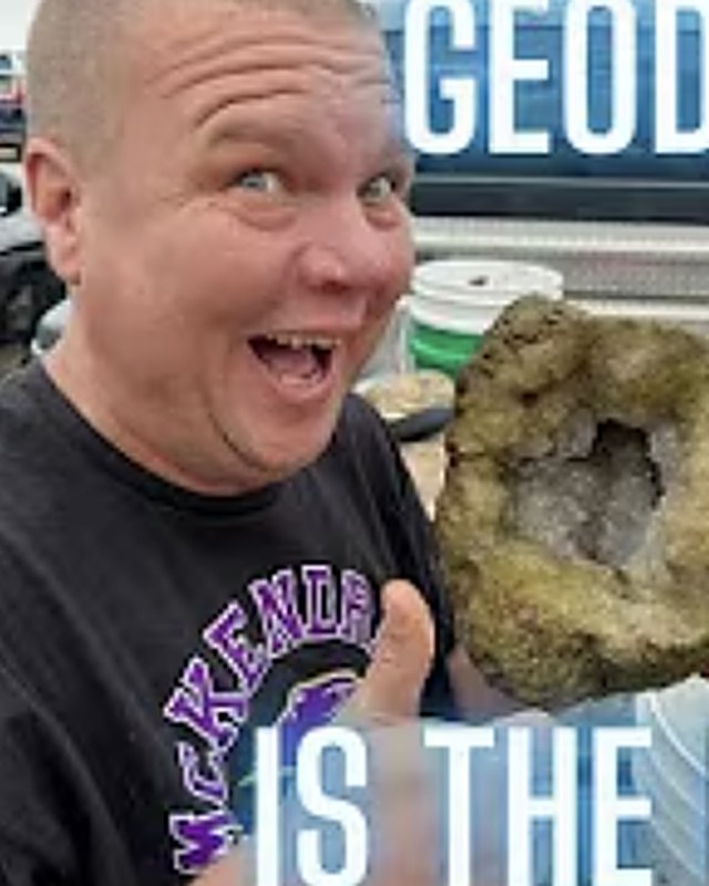 A man grinning and giving a thumbs up beside a split geode lined with quartz crystals.