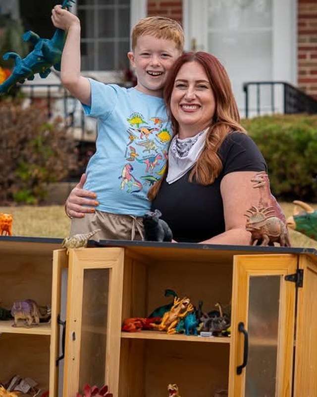A boy and his mother behind a cabinet of toy dinosaurs on a front lawn, under a hand lettered Dino Trade sign.