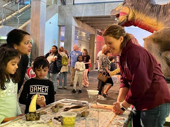 An SLPLS volunteer showing fossils to two children at the Saint Louis Science Center, beneath an animatronic Tyrannosaurus.