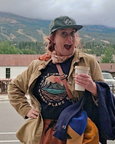 Maggie Mundwiller on a street corner below the mountains, mouth open in mock surprise, holding a coffee.