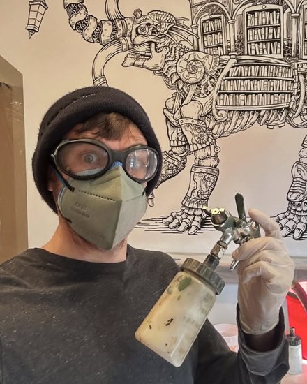 Patrick Weck in goggles and a mask holding an airbrush, in front of his mural of a mechanical elephant carrying a library.